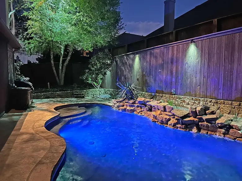 Free-form pool in Colleyville, Texas with a lit rock waterfall and downlights washing the fence line