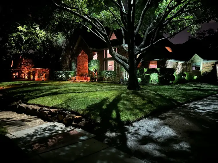 Brick home in Keller, Texas after dark with a grazed stone entry surround and a lit oak canopy throwing a shadow fan across the front lawn