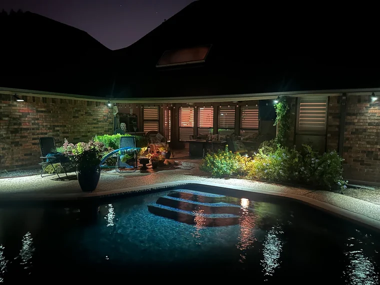 Pool courtyard in Keller, Texas lit low and warm after dark, with washed brick, string lights over the covered patio and planting beds picked out individually